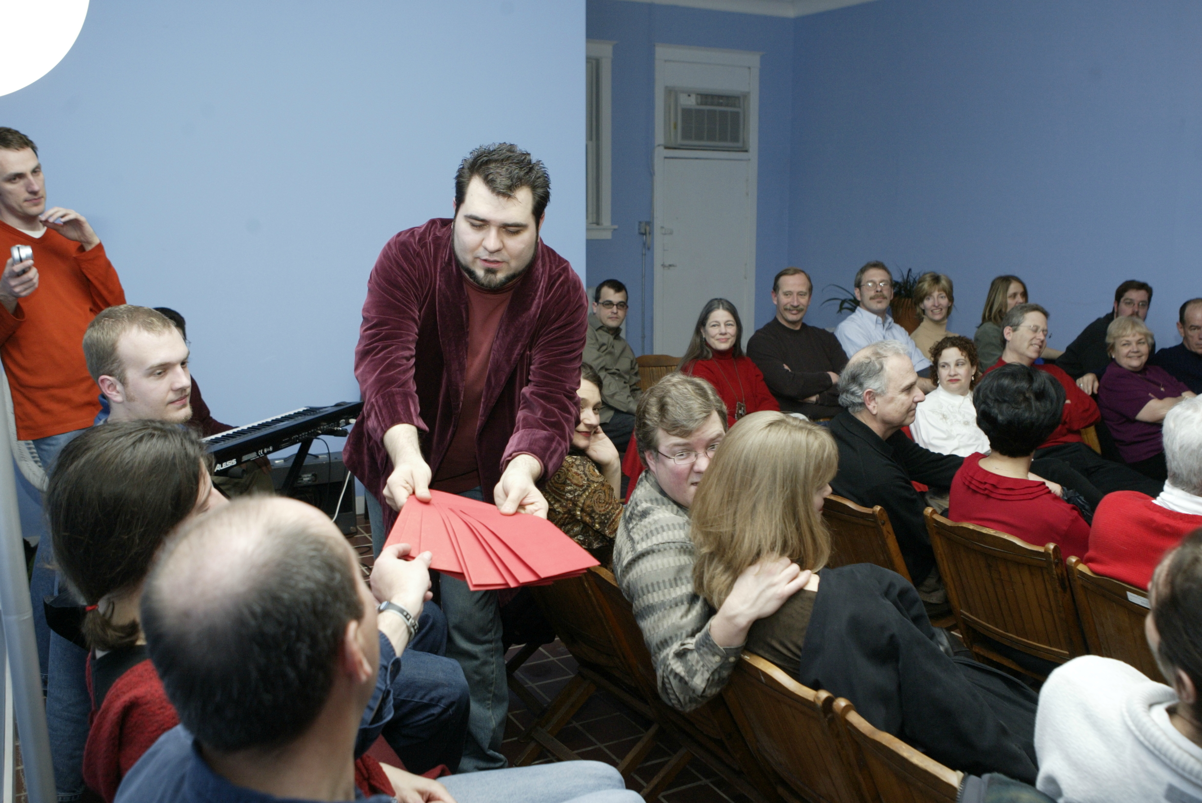 Man dressed in red is handing red envelopes to crowd for selection for improv game. 